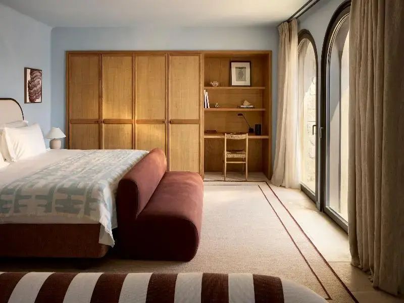 Heller Schlafbereich mit Holzschrank, Schreibtisch und Bett mit rötlicher Sitzbank.