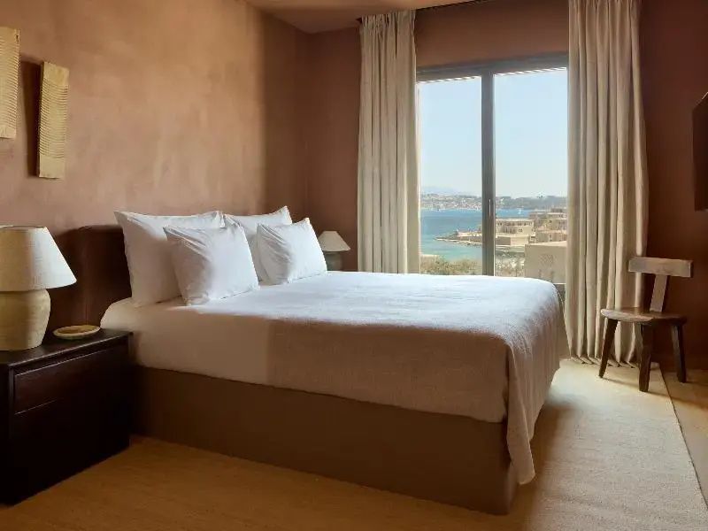 Doppelbett in hellem Zimmer mit Blick durch das Fenster auf das Meer und die Küste.