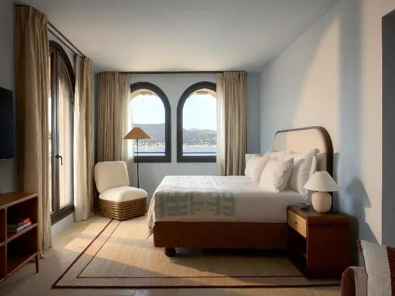 Hell eingerichtetes Schlafzimmer mit Doppelbett, Sessel und zwei Rundbogenfenstern mit Meerblick.