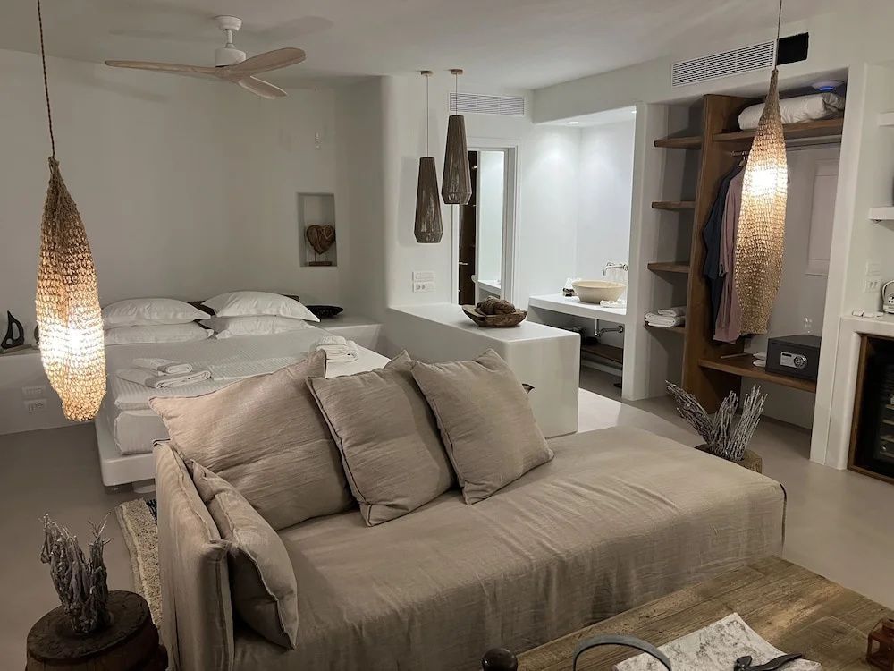 Hell eingerichtetes Hotelzimmer mit Couch im Vordergrund, Doppelbett und Deckenventilator.