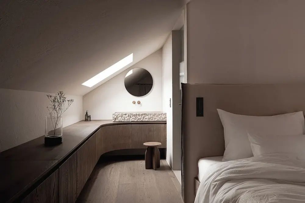 Hotelzimmer unter der Dachschräge mit Holzmöbeln, Steinwaschbecken, rundem Spiegel und einem Dachfenster.
