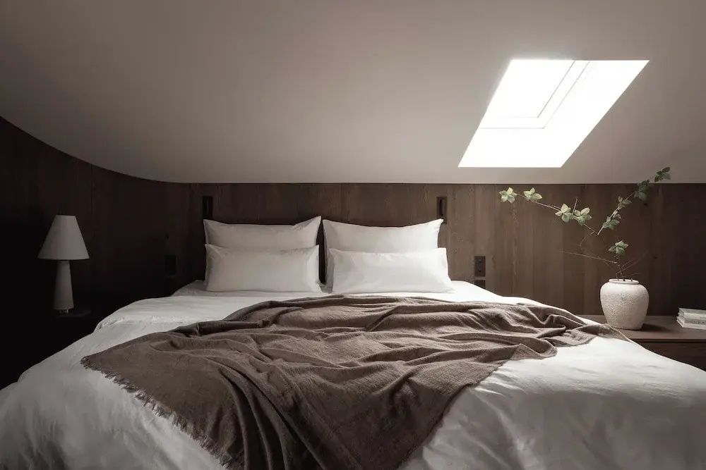 Doppelbett mit weißer Bettwäsche und brauner Tagesdecke unter einem Dachfenster.