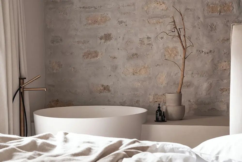 Blick über das Bett auf eine runde weiße Badewanne vor einer beleuchteten Wand aus Naturstein.