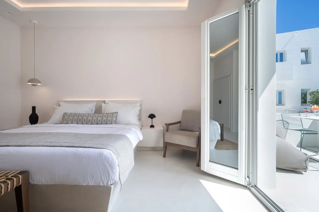 Helles Schlafzimmer mit Doppelbett, weißer Bettwäsche, Sessel und geöffneter Balkontür.