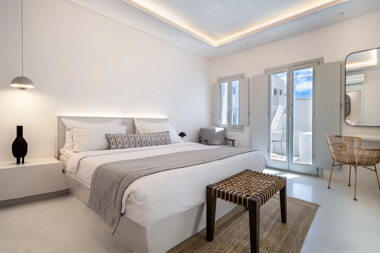 Weiß gestaltetes Schlafzimmer mit großem Doppelbett, Holzbank, Teppich und Fenstertüren.