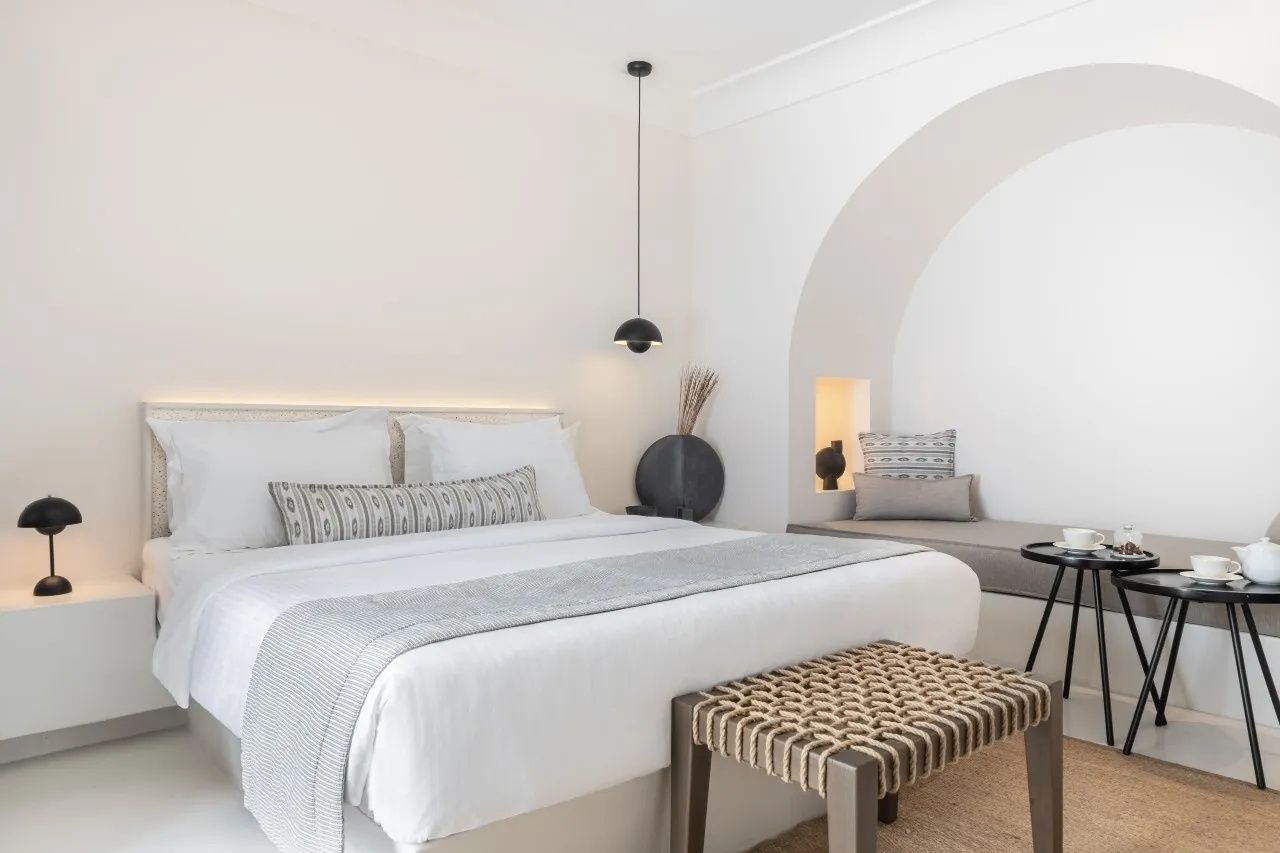 Weiß eingerichtetes Schlafzimmer mit Doppelbett, Flechtbank und einer gemauerten Sitznische in Rundbogenform.