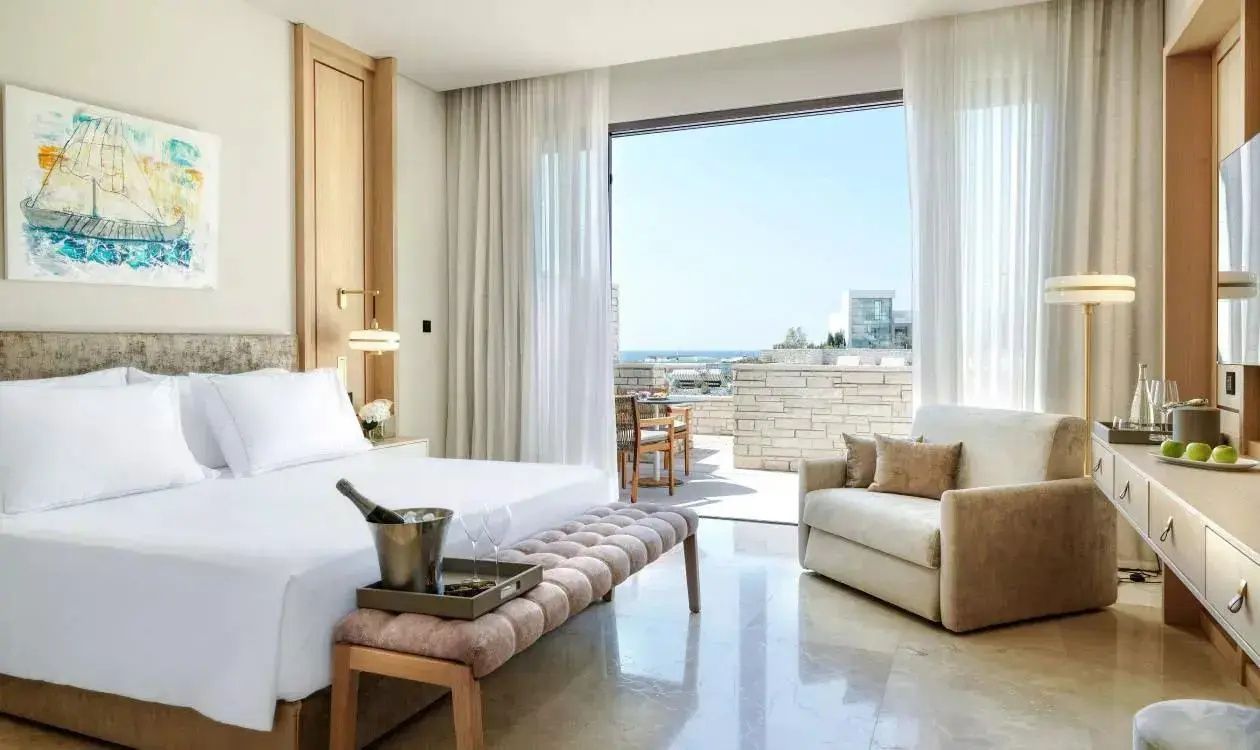 Helles Hotelzimmer mit Doppelbett, Polsterbank, Sessel und Zugang zur Terrasse mit Meerblick.