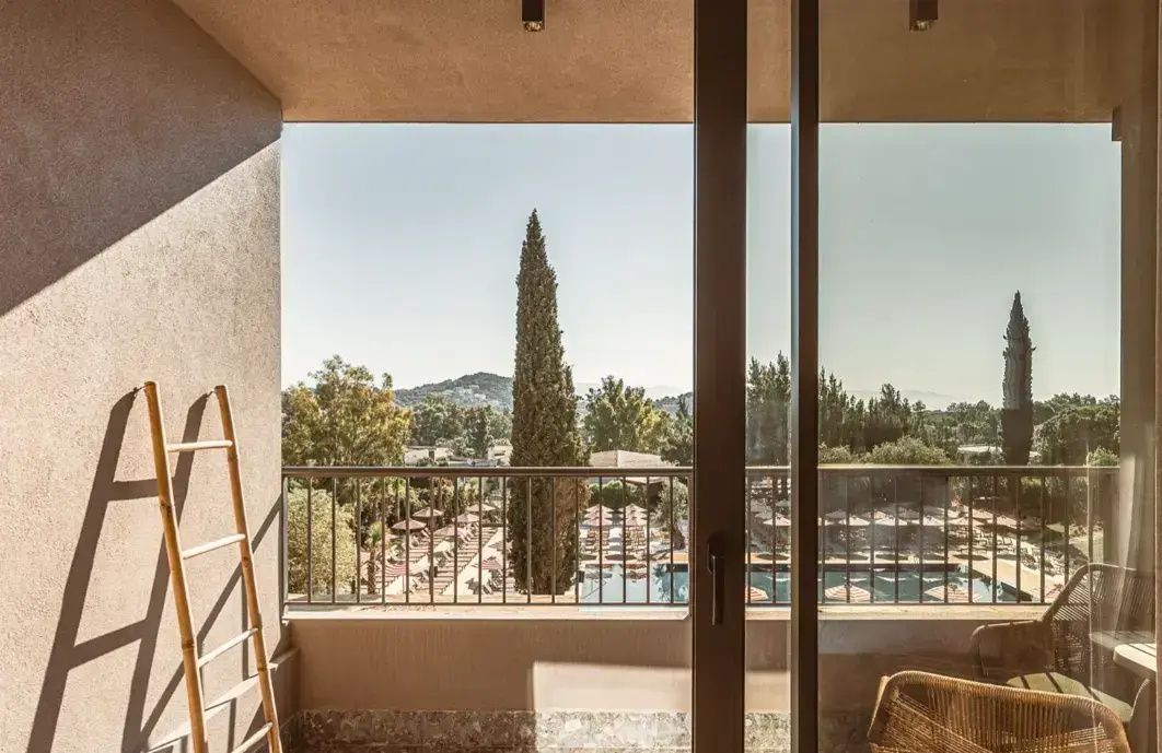 Blick vom überdachten Balkon auf eine grüne Zypresse, den Hotelpool und hügelige Landschaft.
