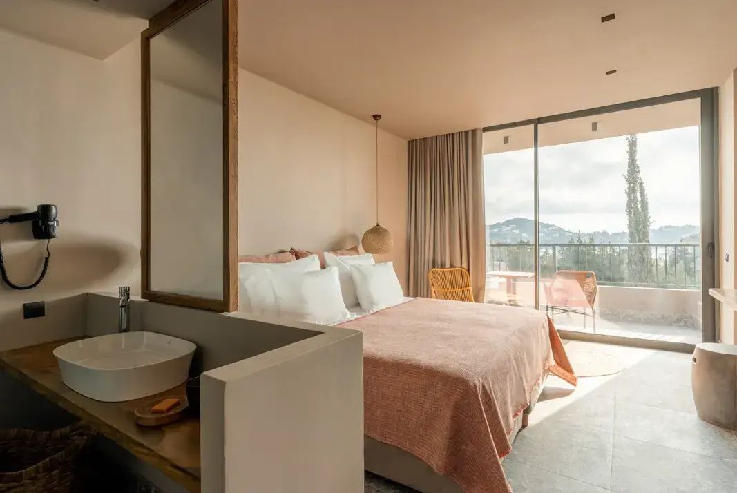 Offenes Hotelzimmer mit großem Doppelbett, Waschtisch und Verglasung zum Balkon.