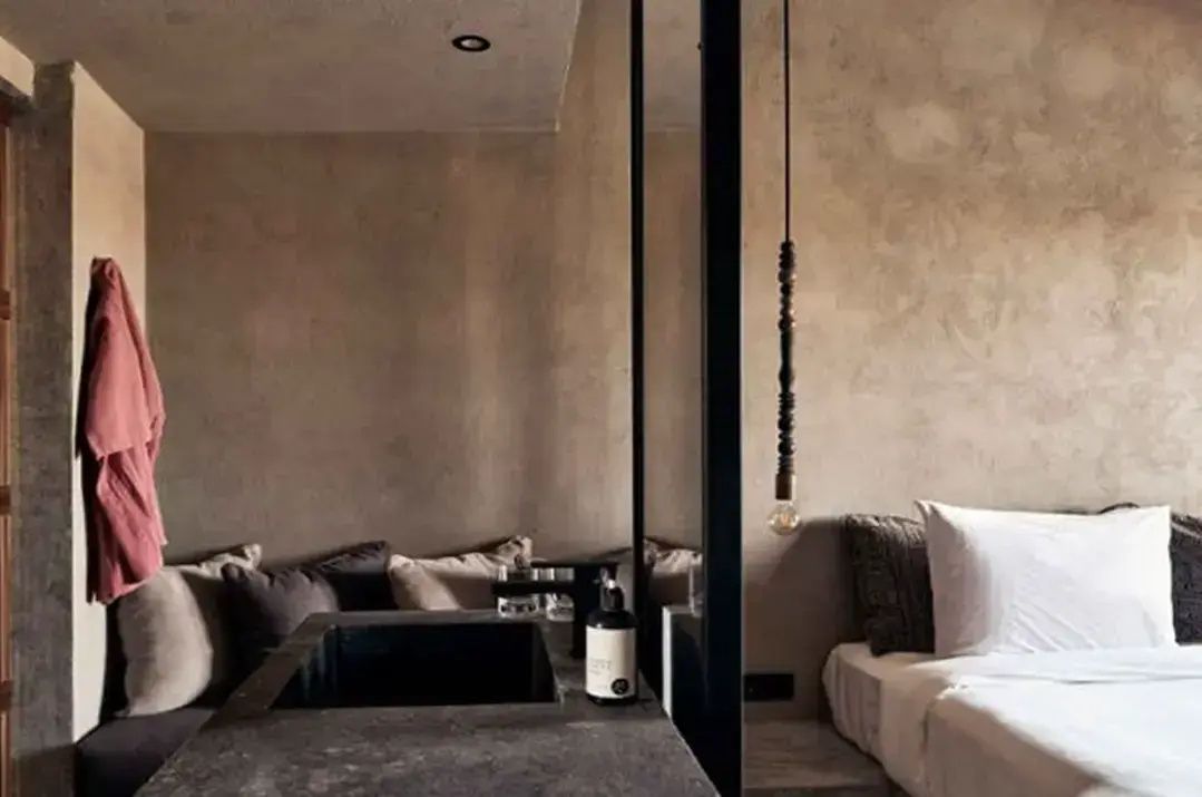 Waschtisch aus Beton im Hotelzimmer neben einer Sitzecke und dem Bett im Hintergrund.