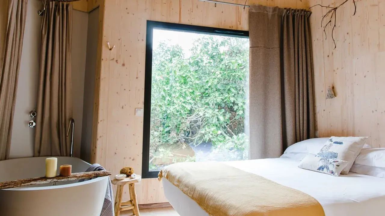 Hotelzimmer mit Holzverkleidung, Doppelbett, freistehender Badewanne und großem Fenster ins Grüne.