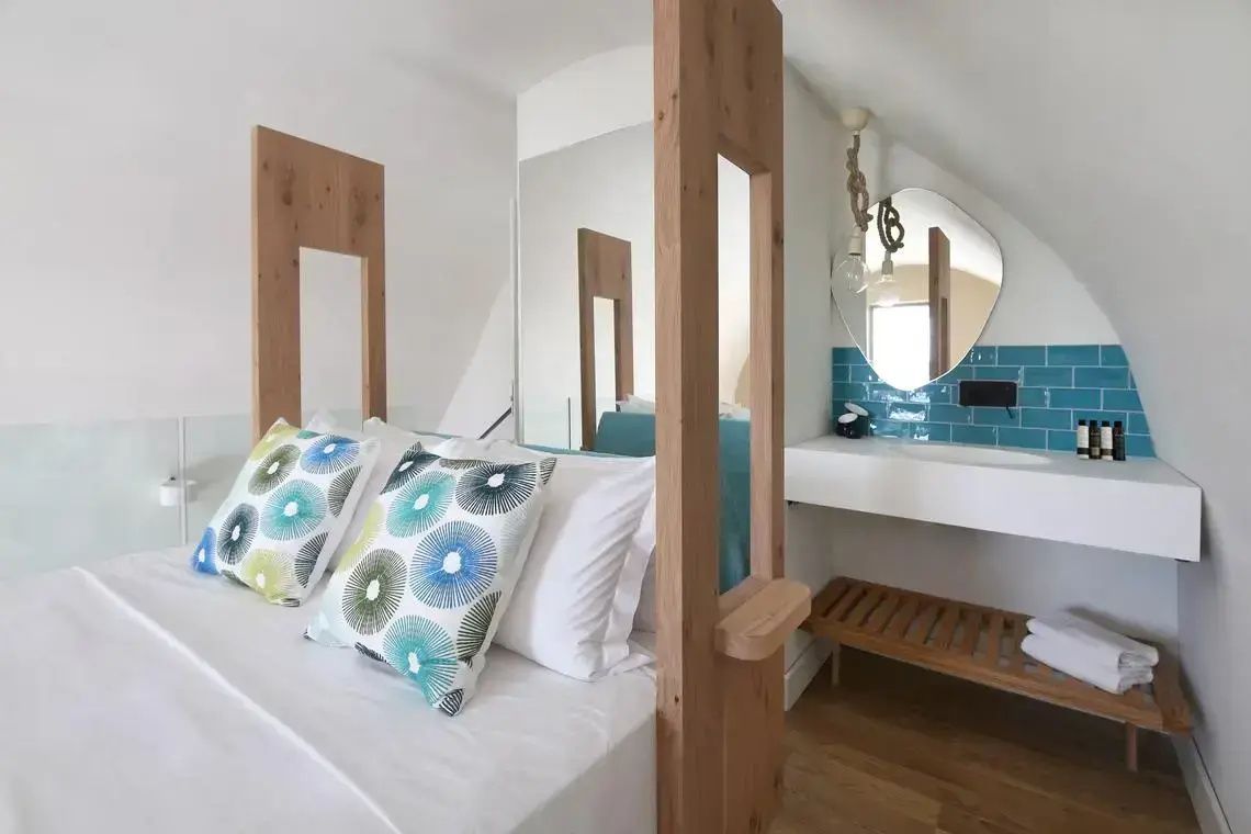 Doppelbett mit Kissen, eine Holzabtrennung und ein sichtbarer Waschtisch mit blauen Fliesen.