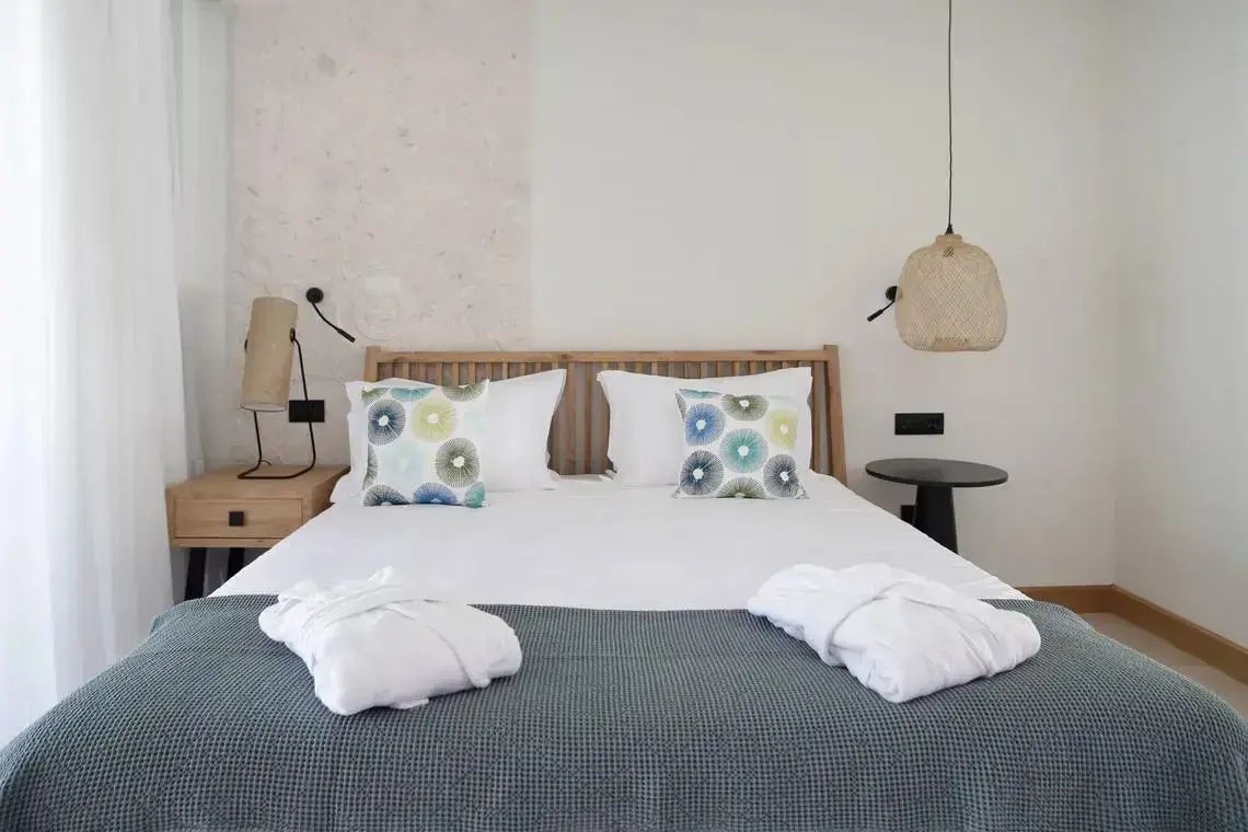 Doppelbett mit weißer Bettwäsche, gemusterten Kissen und zwei Bademänteln.