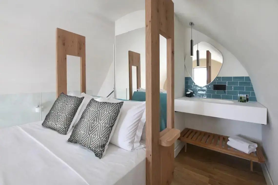 Doppelbett mit grauen Kissen, eine Holzabtrennung sowie ein angrenzendes Waschbecken.