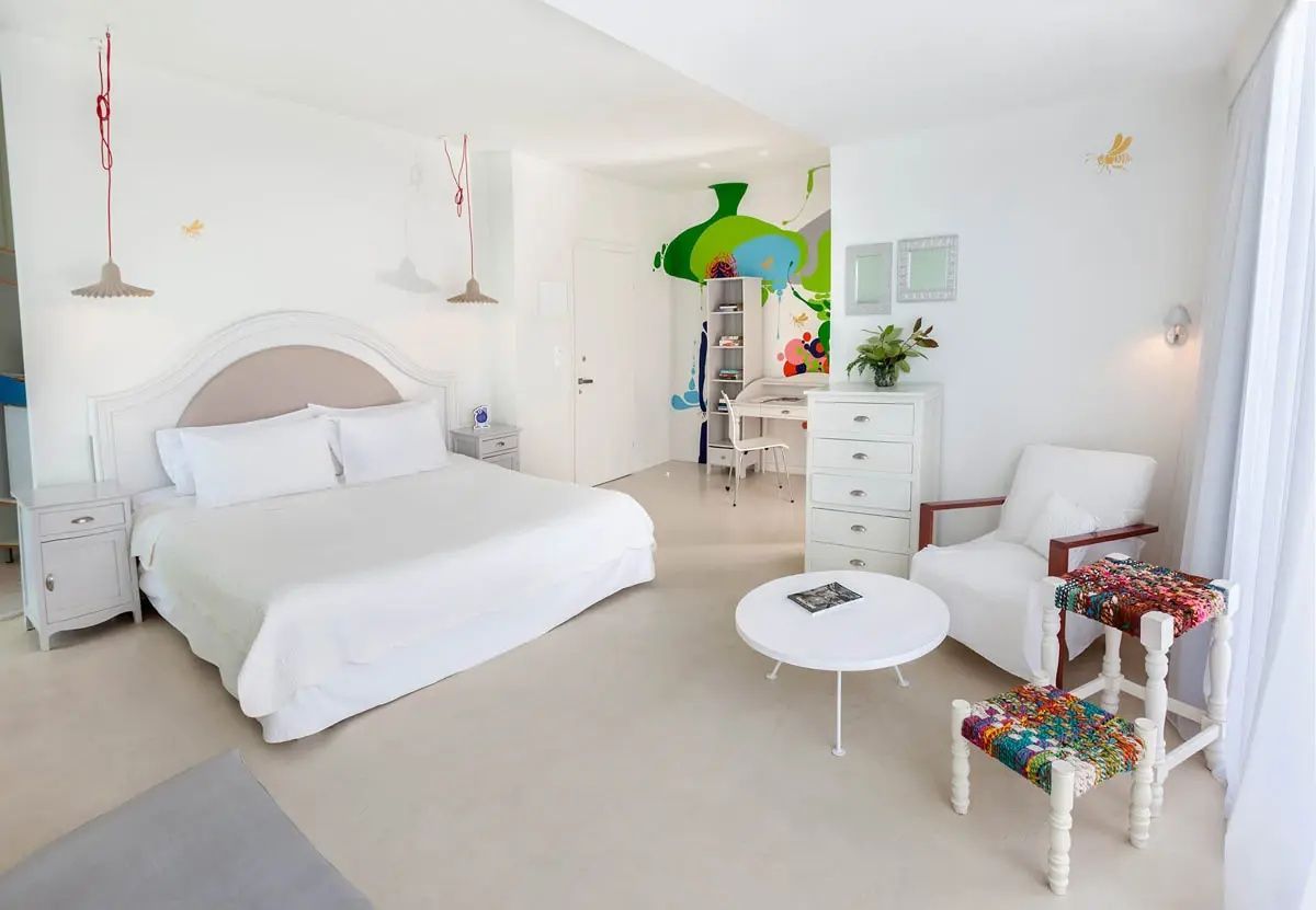 Geräumiges weißes Zimmer mit großem Bett, Kommode und bunten Kunstakzenten an der Wand.