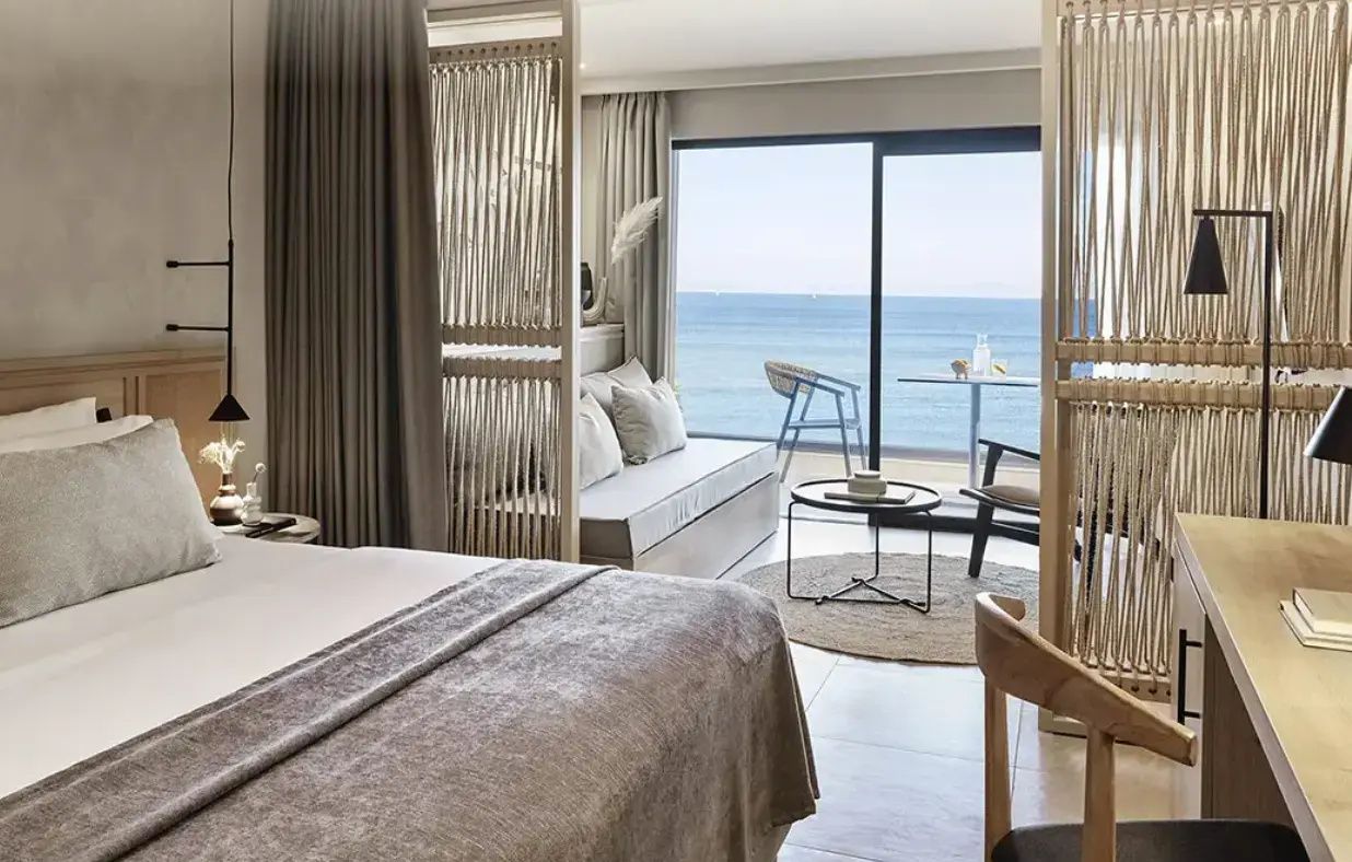 Hotelzimmer mit Sitzbereich und Blick durch die Glasfront auf die Terrasse und das Meer.