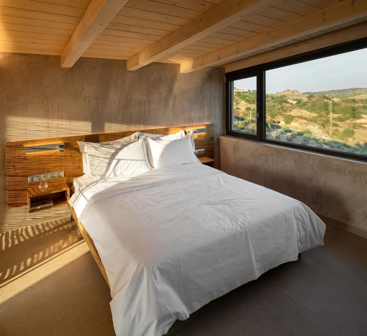 Modernes Schlafzimmer mit Holzkopfteil, weißem Doppelbett und Fenster ins Grüne.