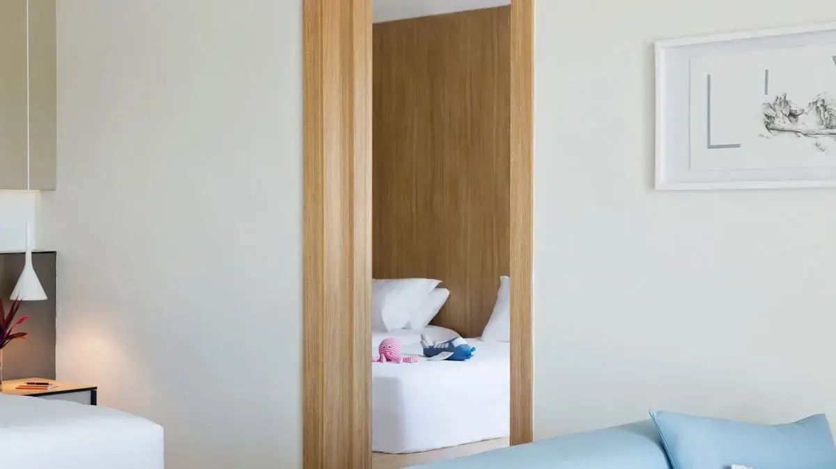 Blick durch einen hölzernen Türrahmen auf ein weiß bezogenes Bett im Hotelzimmer.