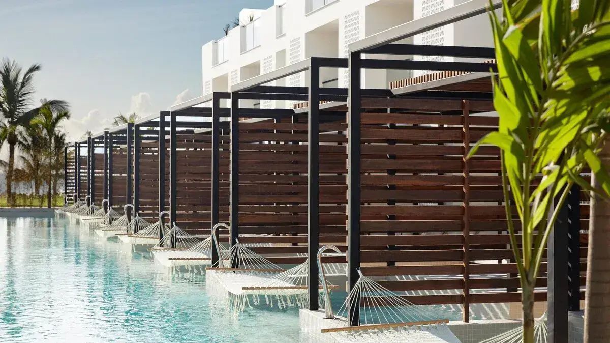 Hängematten über dem Poolwasser vor hölzernen Sichtschutzwänden und weißer Hotelfassade.