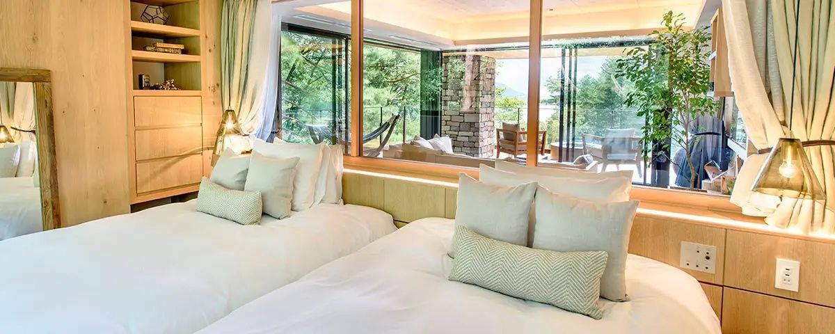 Schlafbereich mit weiß bezogenen Betten und Fensterscheibe zum überdachten Balkon.