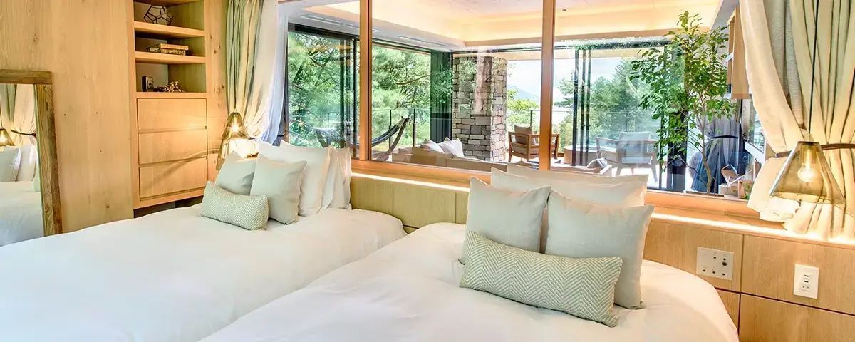 Betten mit hellen Kissen in einem Hotelzimmer mit Blick auf die Glasfront zur Terrasse.