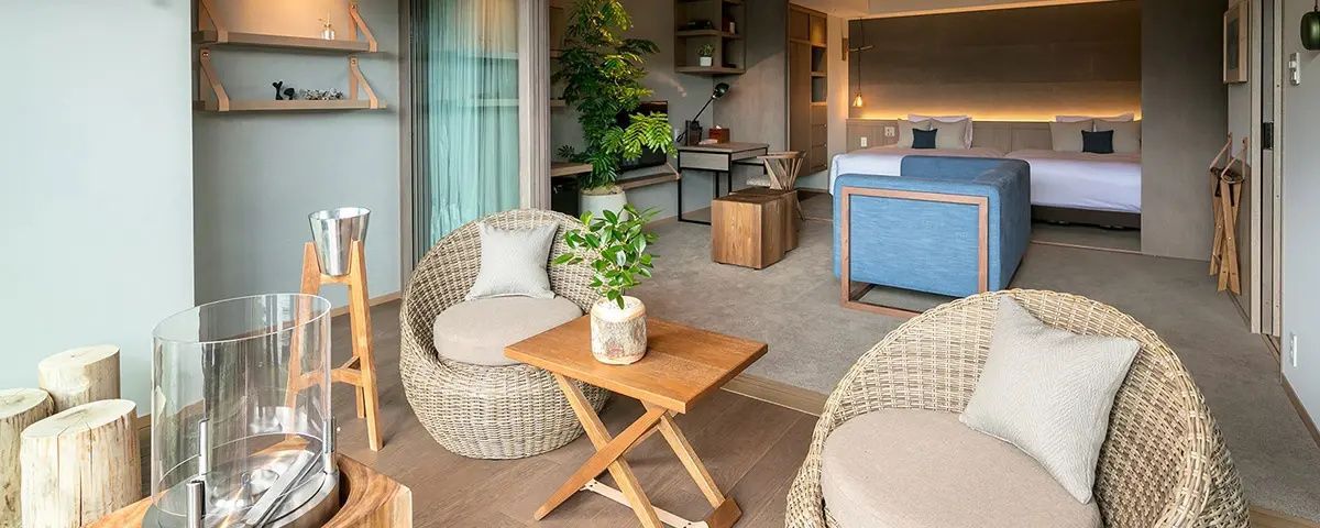 Sitzbereich einer Suite mit Flechtsesseln, Holztisch, großer Zimmerpflanze und Bett im Hintergrund.