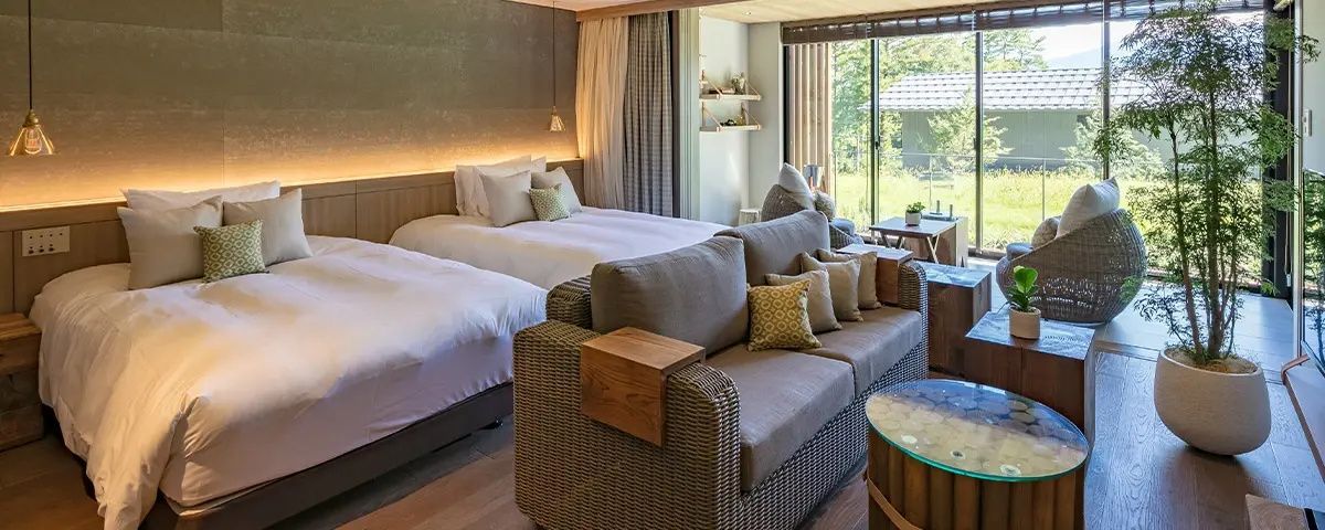 Hotelzimmer mit zwei großen Betten, einem geflochtenen Sofa und warmem Licht an der Wand.