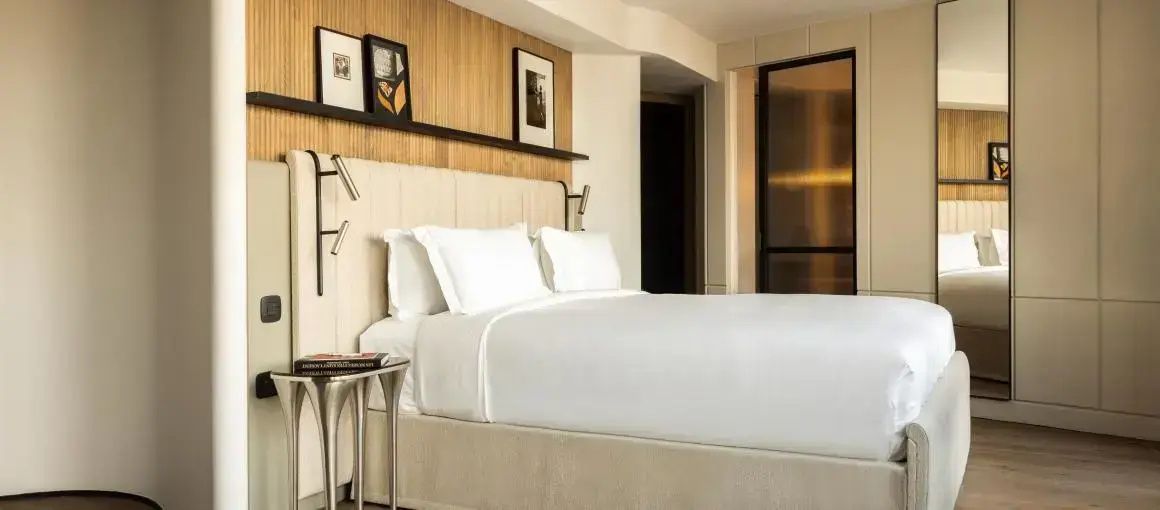 Doppelbett mit weißer Bettwäsche, gepolstertem Kopfteil und Wandregal im Hotelzimmer.