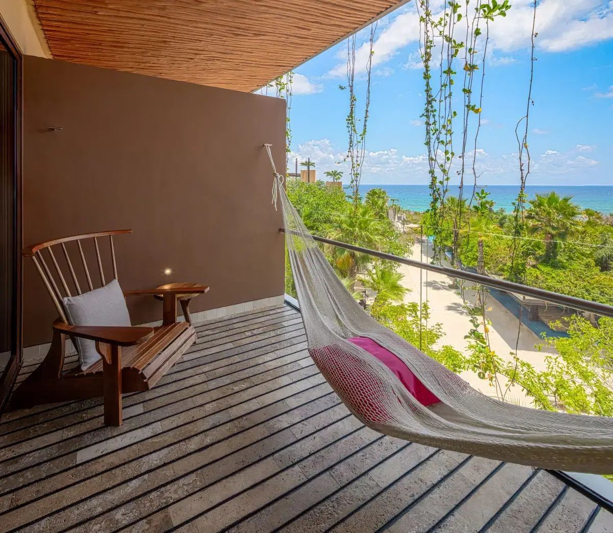 Balkon mit Hängematte und Holzstuhl sowie Blick auf grüne Pflanzen und das Meer.