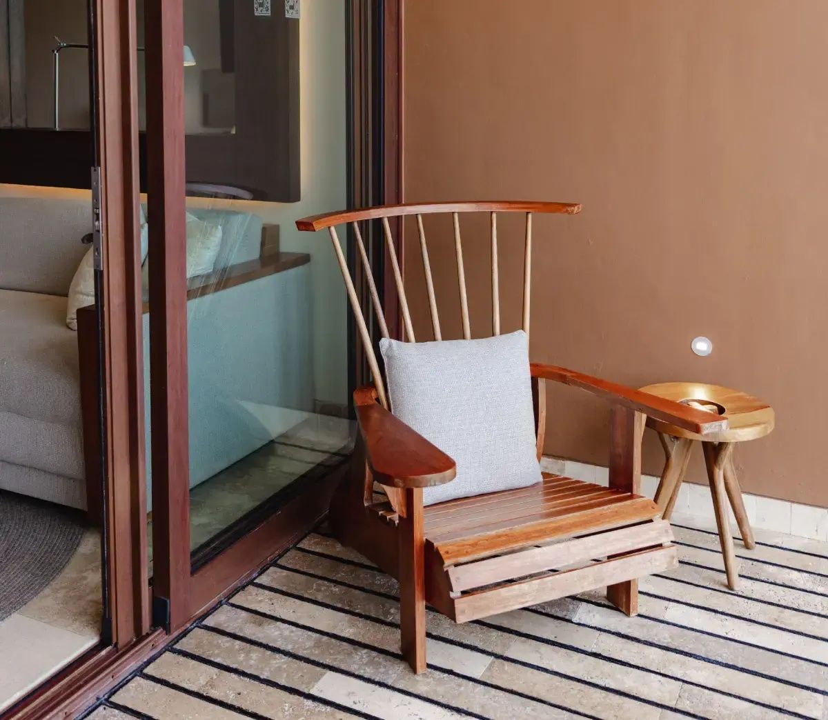 Ein Holzsessel mit hellem Kissen auf einer Terrasse vor den Schiebetüren zum Zimmer.