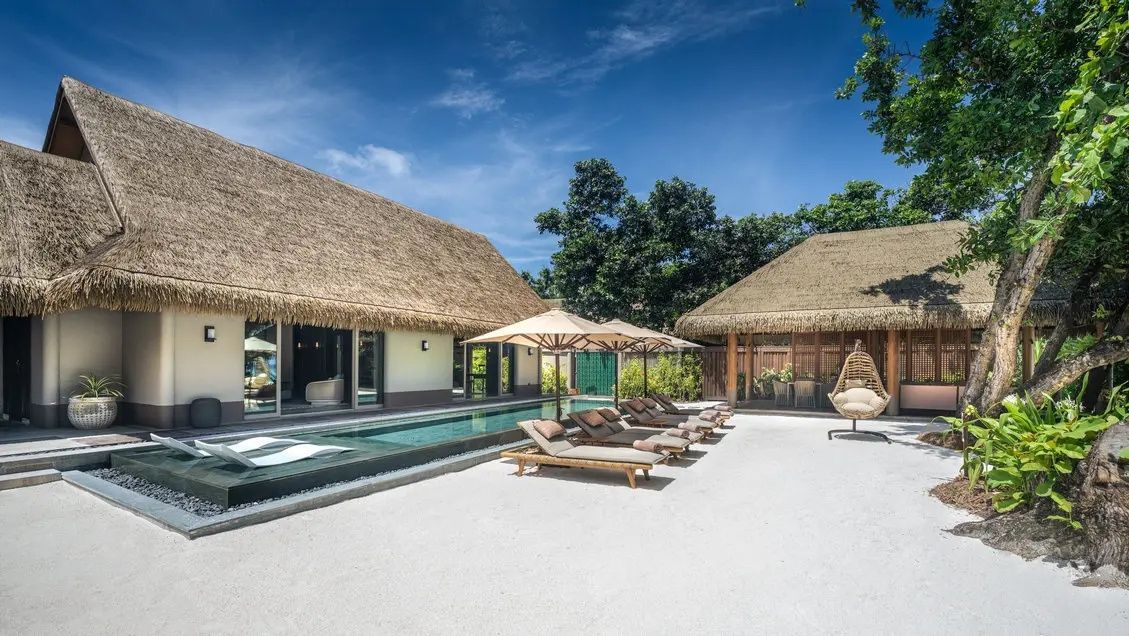 Reetgedeckte Villa mit großem Außenpool, Terrasse und Liegestühlen auf Sand.