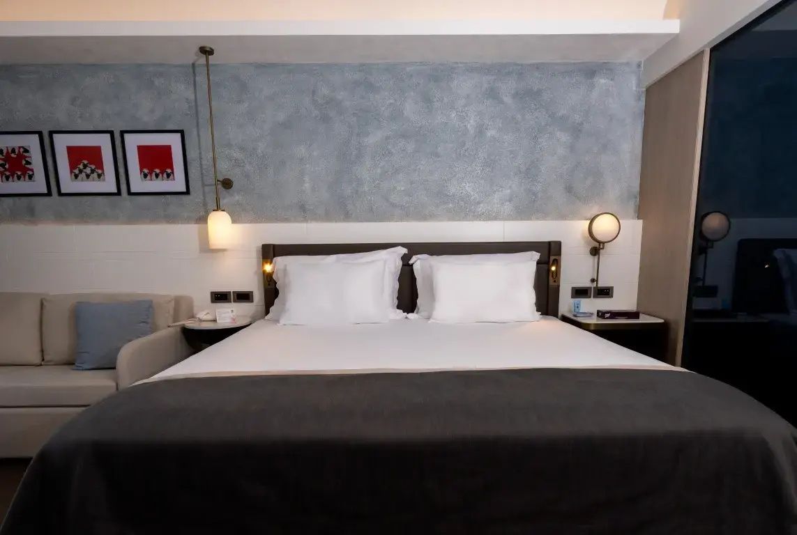 Großes Doppelbett mit weißen Kissen und grauer Decke vor einer getönten Wand.
