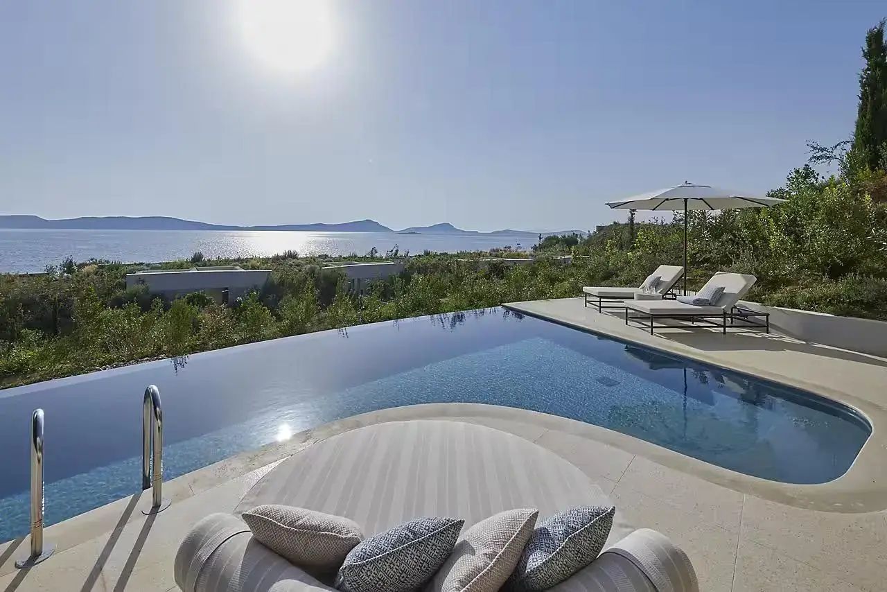 Infinity-Pool mit Sonnenliegen und freiem Blick auf das Meer sowie die Berge bei hellem Sonnenschein.