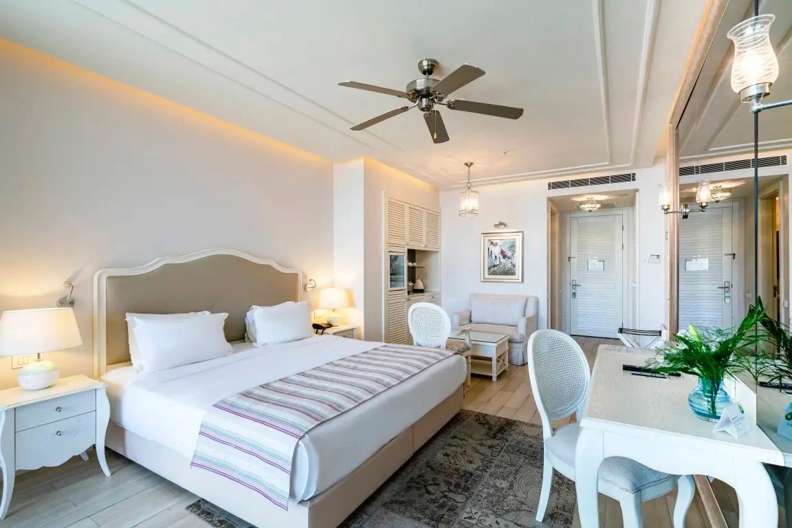 Hell eingerichtetes Hotelzimmer mit Doppelbett, Deckenventilator und Schreibtisch.