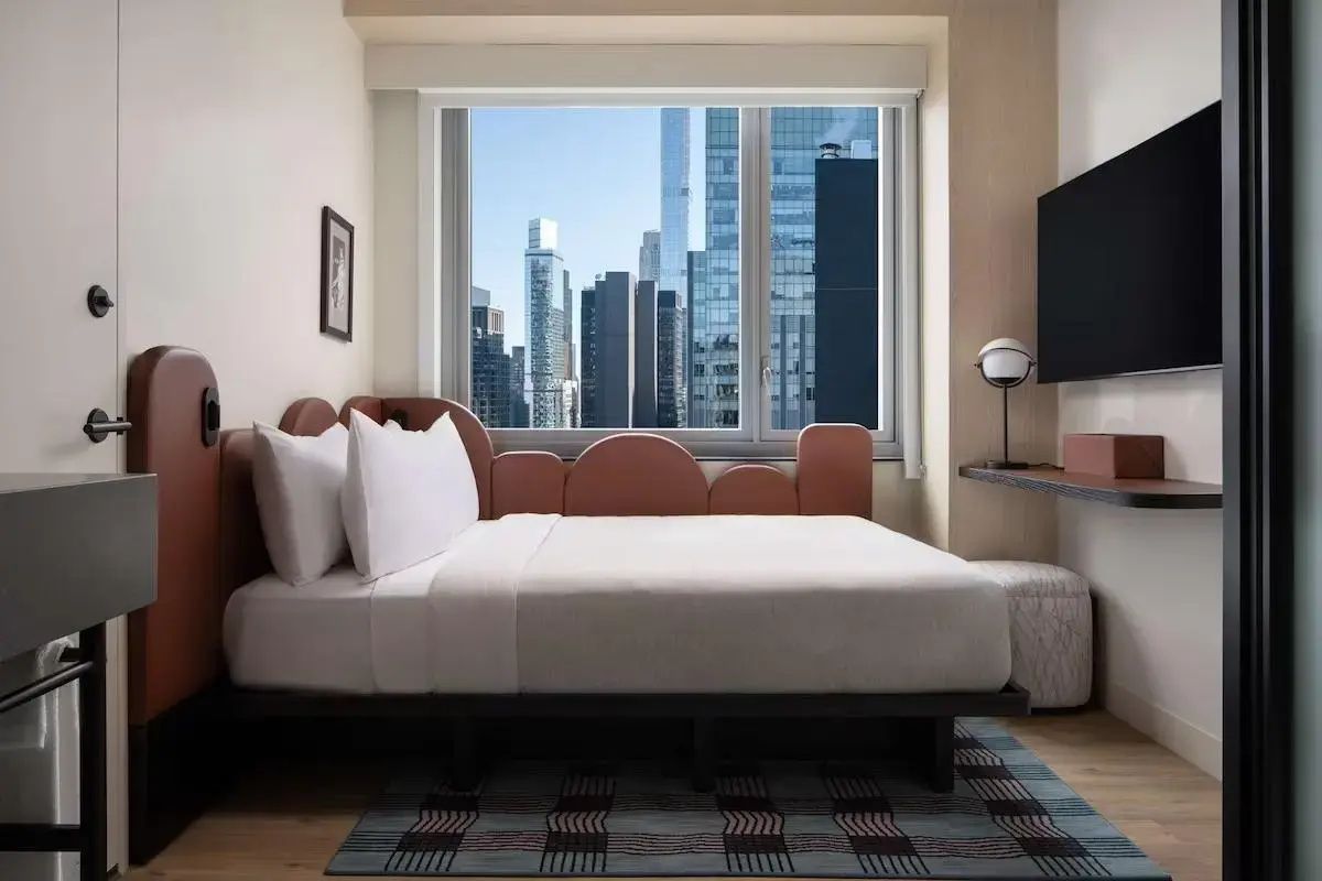 Heller Schlafraum mit weiß bezogenem Bett, Teppich und Ausblick auf die Skyline der Stadt.