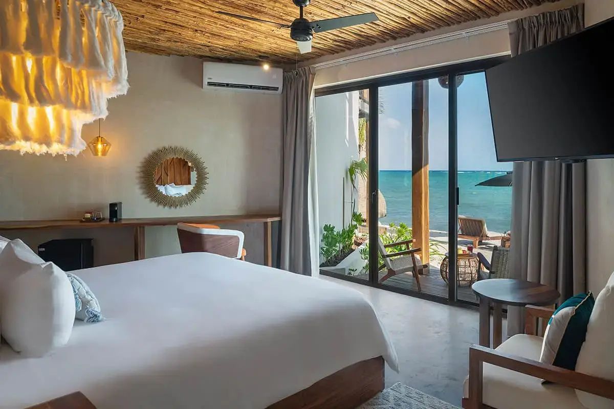 Helles Hotelzimmer mit weißem Doppelbett und Blick durch die Glastür auf das Meer.