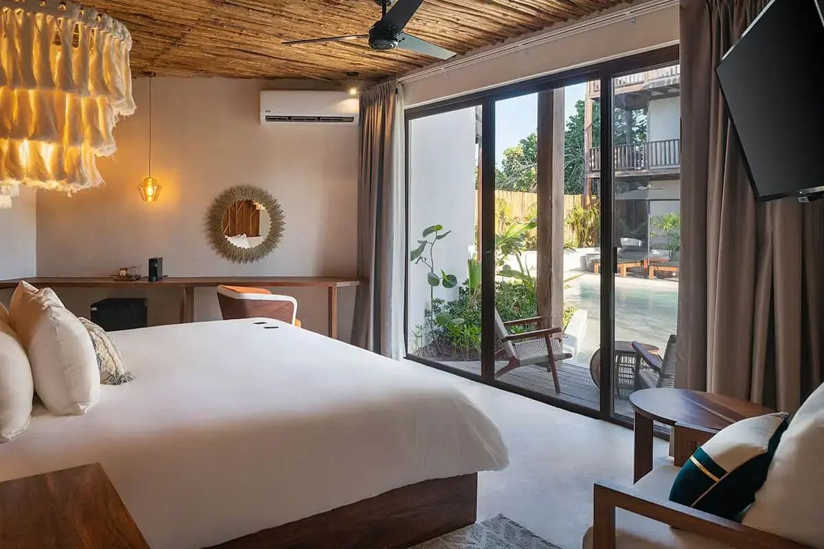 Helles Hotelzimmer mit Doppelbett, Holzdecke und großem Glasfenster mit Zugang zum Poolbereich.