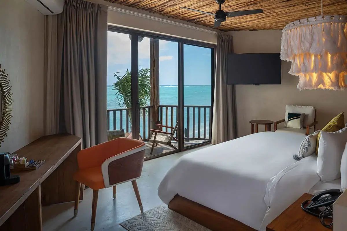 Hotelzimmer mit weißem Bett, Holzdecke und Balkontür mit Blick auf das türkisfarbene Meer.
