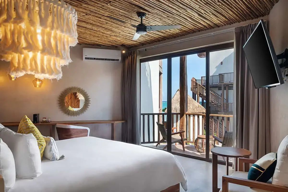 Hotelzimmer mit weiß bezogenem Bett, Bambusdecke und Balkontür mit Blick nach draußen.