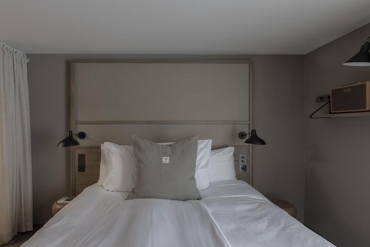 Doppelbett mit weißen Kissen, grauer Decke und Wandlampen im Hotelzimmer.