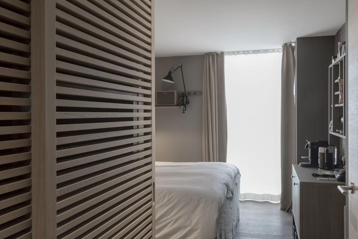 Blick durch eine Holzlamellentür auf ein helles Bett am Fenster.