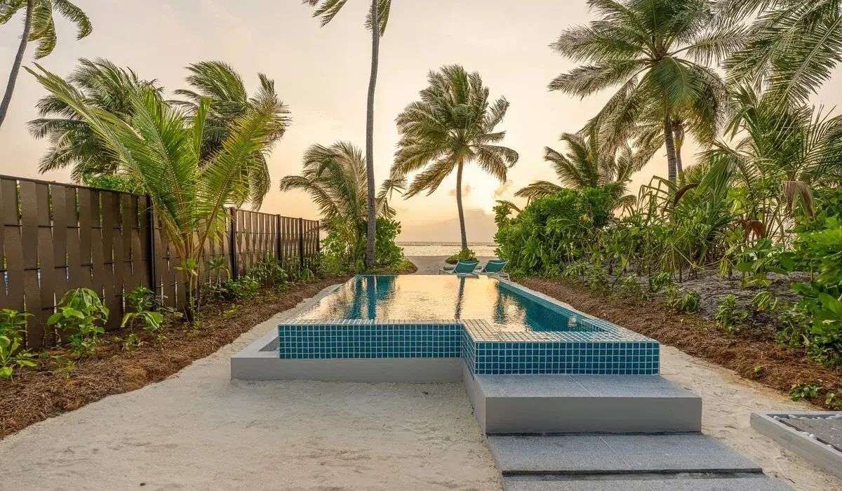 Rechteckiger Pool auf einem Sandstrand, umgeben von Palmen und tropischer Vegetation.
