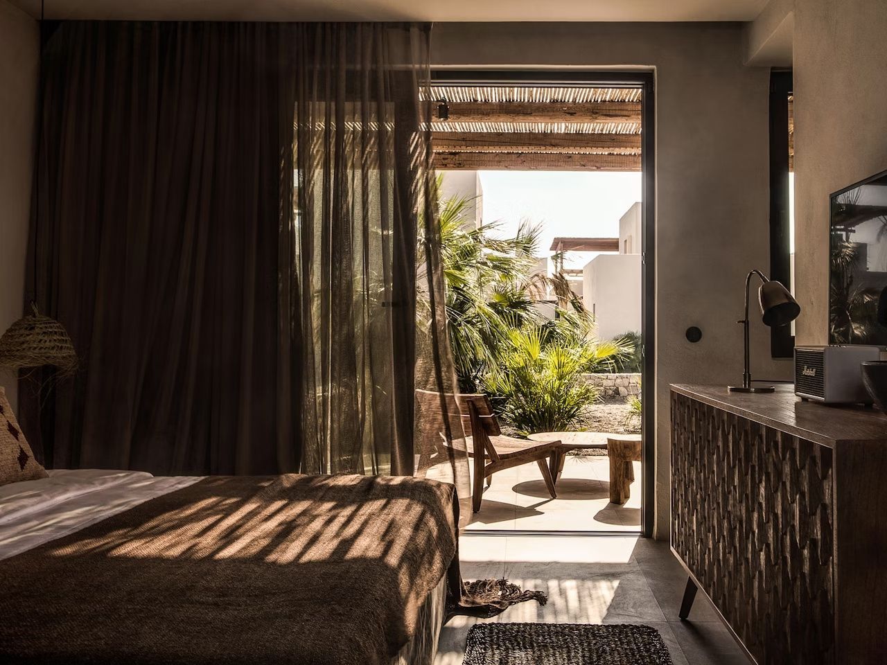 Blick aus dem abgedunkelten Hotelzimmer auf eine sonnige Terrasse mit Holzmöbeln und grünen Pflanzen.