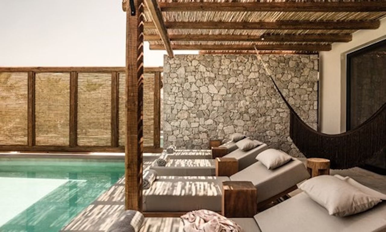 Sonnenliegen aus Holz unter einem Pergoladach direkt am Pool vor einer Steinwand.
