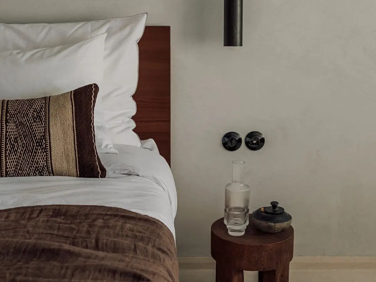 Ein Bett mit Dekokissen neben einem kleinen Holztisch mit Wasserflasche und Glas.