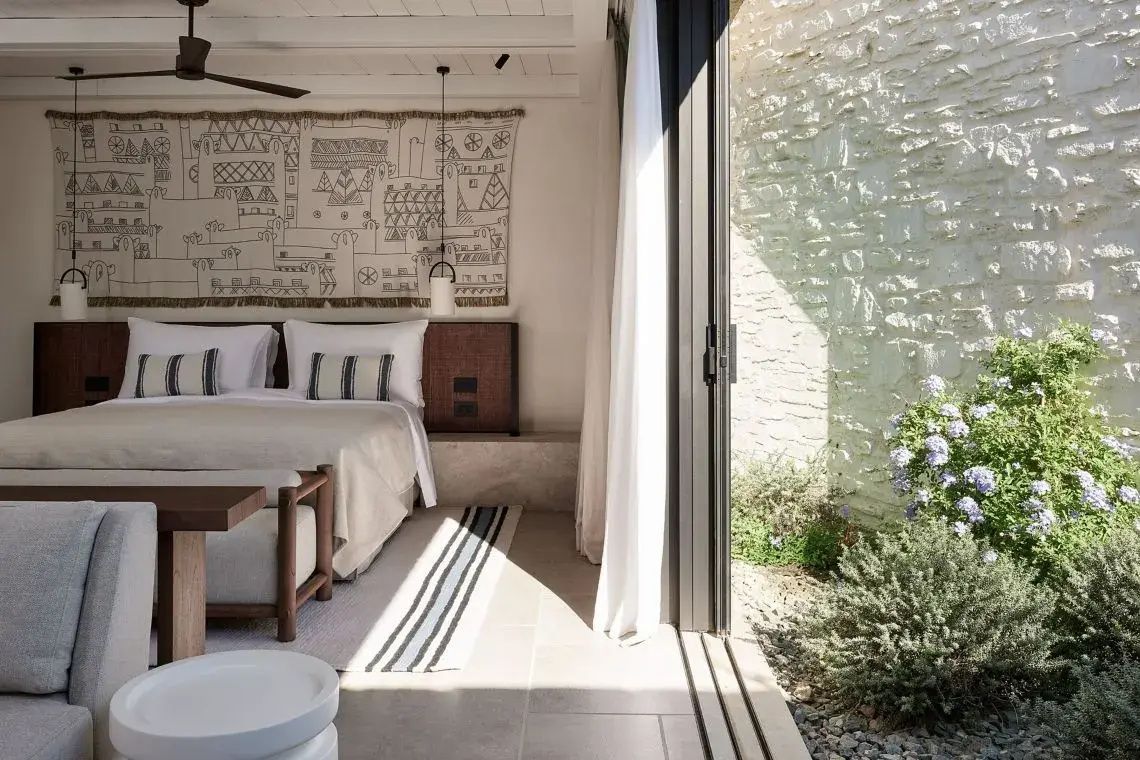 Helles Hotelzimmer mit Doppelbett, Wandteppich und geöffneter Terrassentür zum Garten.
