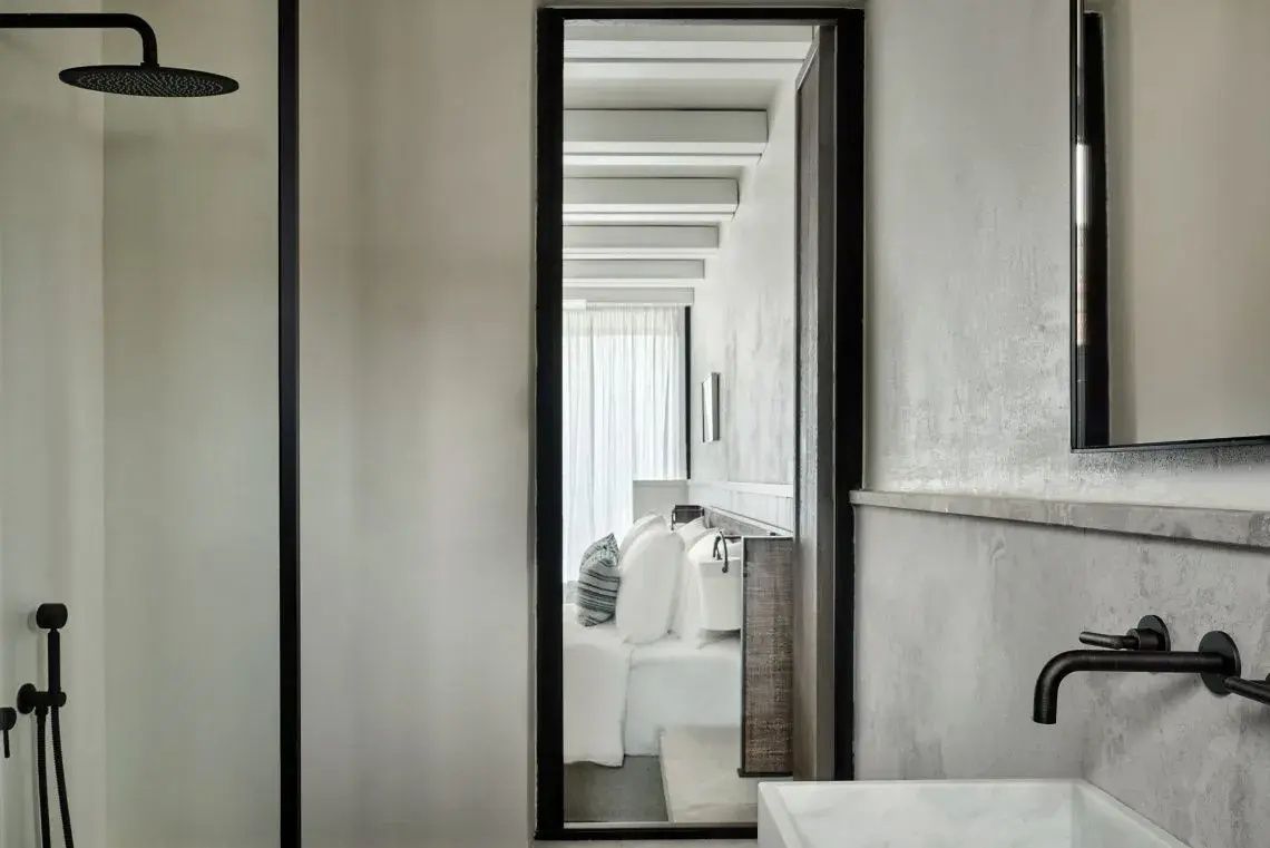 Blick aus dem Badezimmer in ein helles Schlafzimmer mit Holzbalken an der Decke.