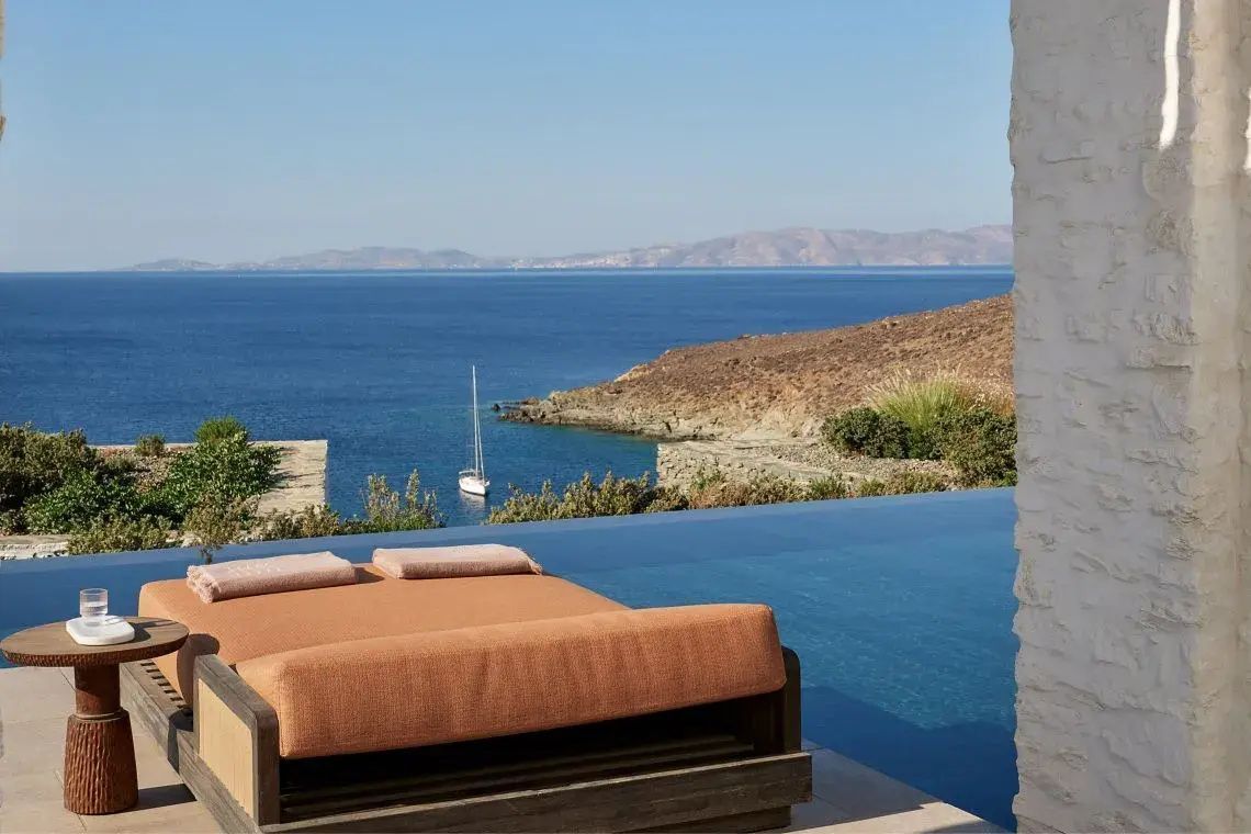 Terrassen-Tagesbett an einem Infinity-Pool mit Blick auf das blaue Meer und hügelige Küstenlandschaft.