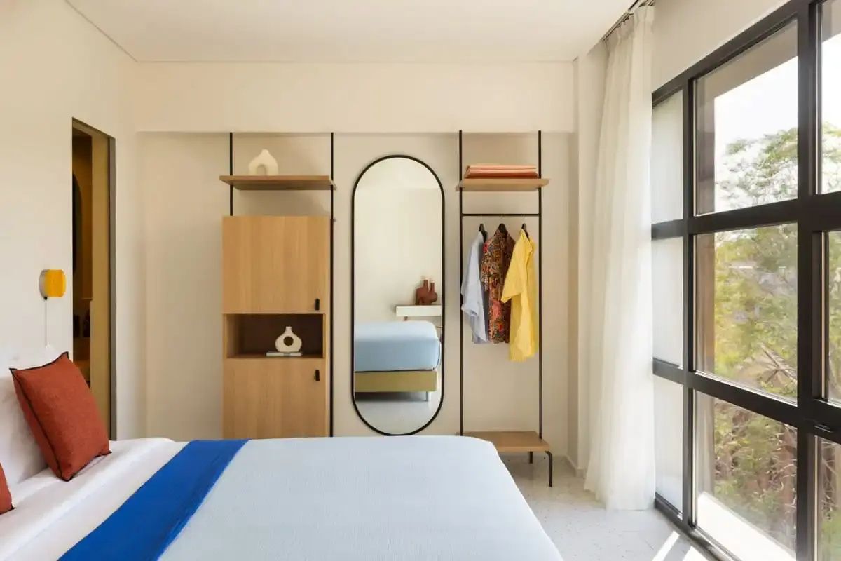 Hell eingerichtetes Hotelzimmer mit Doppelbett, einem ovalen Wandspiegel und offenem Kleiderschrank.