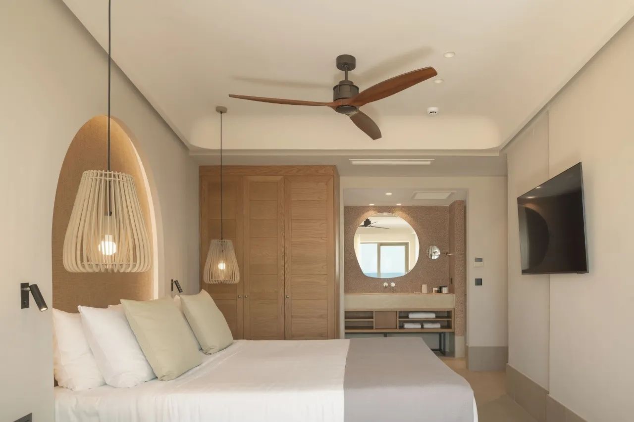 Hell eingerichtetes Hotelzimmer mit Doppelbett, Deckenventilator aus Holz und Blick ins Bad.